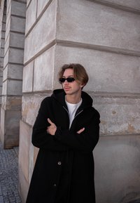 Črn volnen plašč z zadrgo na ovratniku, oblečen čez belo srajco. Črne pravokotne son�čna očala in ozadje iz teksturirane kamnite stene.