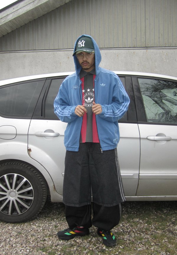 Hombre con sudadera azul Adidas sobre camiseta gráfica, pantalones cortos negros anchos, zapatillas multicolor y gorra de New York Yankees, está junto a un coche plateado.