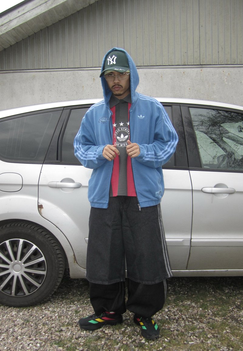 Un homme porte un sweat à capuche Adidas bleu sur un t-shirt graphique, un short noir large, des baskets multicolores et une casquette des New York Yankees, il se tient à côté d'une voiture argentée.