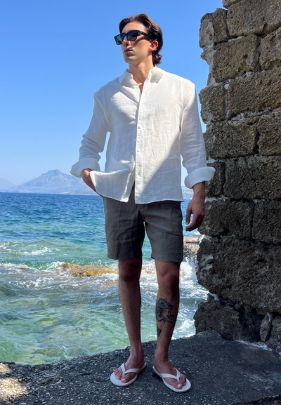 Camisa de lino blanca, pantalones cortos grises y chanclas blancas que lleva una persona cerca del agua, con un muro de piedra al fondo y montañas a la distancia.