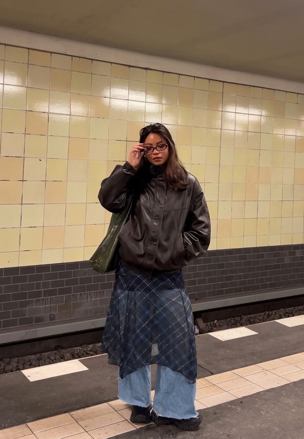 Kvinde justerer briller og står på metrostation iført mørk læderjakke, lagdelt nederdel over brede jeans og bærer en grøn taske.