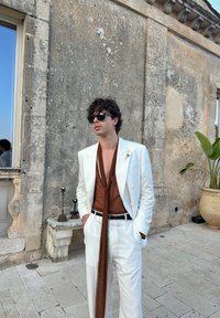 Uomo con capelli ricci che indossa occhiali da sole, un completo bianco e una camicia trasparente marrone con una lunga sciarpa, in piedi contro un muro di pietra invecchiato.