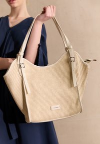 Beige gewebte Tragetasche mit glatten, hellen Lederriemen und einer weichen Textur, versehen mit einem kleinen Markenlabel auf der Vorderseite.