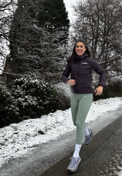 Chaqueta acolchada negra tipo puffer, leggings verde menta, calcetines deportivos blancos y zapatillas de correr grises. Camino nevado y árboles de fondo.