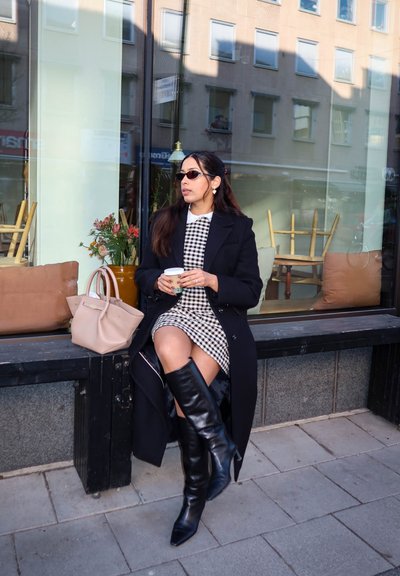 Mujer con abrigo negro, vestido a cuadros y botas hasta la rodilla sentada en un banco frente a una cafetería sosteniendo una taza de café, con gafas de sol y bolso a su lado.