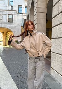 Béžová zkrácená bunda s vysokým límcem a oversized střihem, zdobená velkými černými knoflíky. Nosit�á s světle šedými širokými džíny na dlažební ulici.