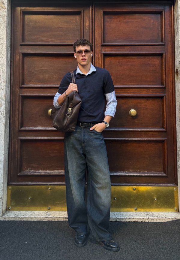 Polo shirt scuro, camicia azzurra a collo sotto, jeans in denim scuro taglia oversize, scarpe nere, tiene in mano una borsa in pelle marrone, in piedi accanto a una porta di legno.