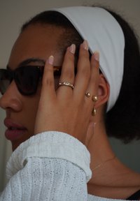 Zonnebril met donkere lenzen, een witte hoofdband en gouden sieraden, waaronder een ring met stenen, pareloorbellen en een gestructureerde witte trui.