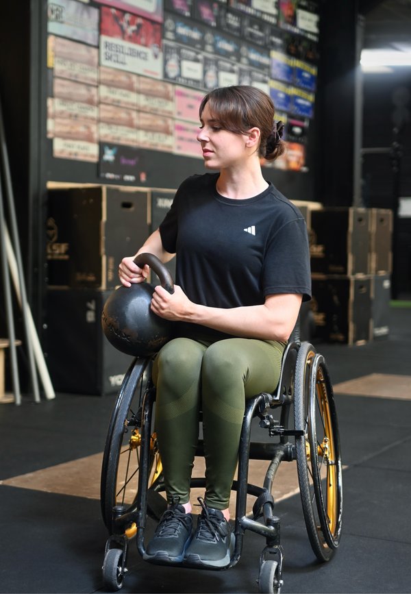 Femme en fauteuil roulant tenant un kettlebell, vêtue d'un t-shirt noir et de leggings verts, faisant de l'exercice dans une salle de sport avec du matériel d'entraînement en arrière-plan.