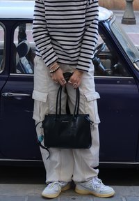 Bolsa de couro preta com alças curtas, logotipo à frente. Camisa de manga longa casual com riscas, calças cargo bege, sapatilhas desportivas com detalhes coloridos.