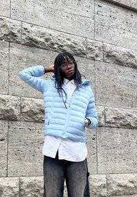 Šviesiai mėlyna, paminkštinta striukė su užtrauktuku priekyje ir gobtuvu, dėvima virš baltos marškinėlių. Yra tekstūruota medžiaga ir laisvas fitas.