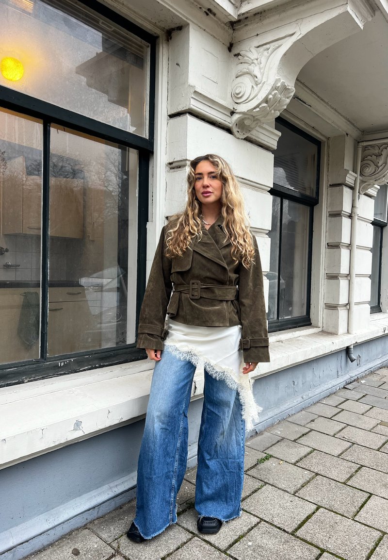 Femme aux longs cheveux bouclés portant une veste olive ceinturée, une jupe asymétrique blanche bordée de dentelle sur un jean bleu à jambes larges, debout sur un trottoir.