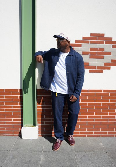 Chaqueta de mezclilla azul y pantalones sobre una camisa blanca, combinados con zapatos burdeos. Una gorra ligera y gafas de sol redondas completan el look.