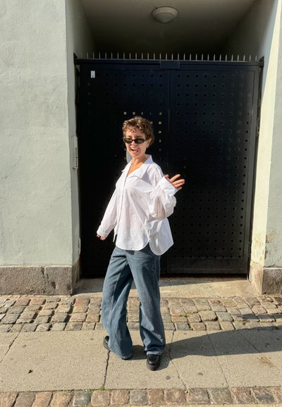 Persona con gafas de sol, una camisa blanca holgada y jeans de pierna ancha, sonriendo y saludando frente a una gran puerta de metal negro.