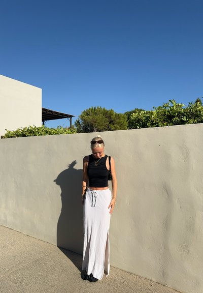 Top corto sin mangas negro, falda maxi blanca con abertura lateral y zapatos oscuros. El fondo presenta una pared texturizada y vegetación bajo un cielo azul despejado.