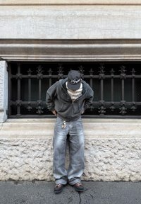 Felpa grigia con cerniera e t-shirt bianca, jeans larghi azzurri, scarpe marroni. Persona in piedi vicino a una grata per finestra in ferro battuto nera.