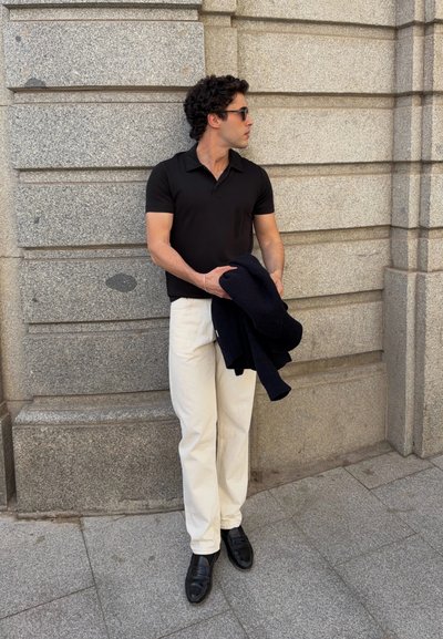 Hombre con camiseta polo negra, pantalones blancos y zapatos negros, sosteniendo una chaqueta oscura doblada, de pie junto a una pared de piedra gris.