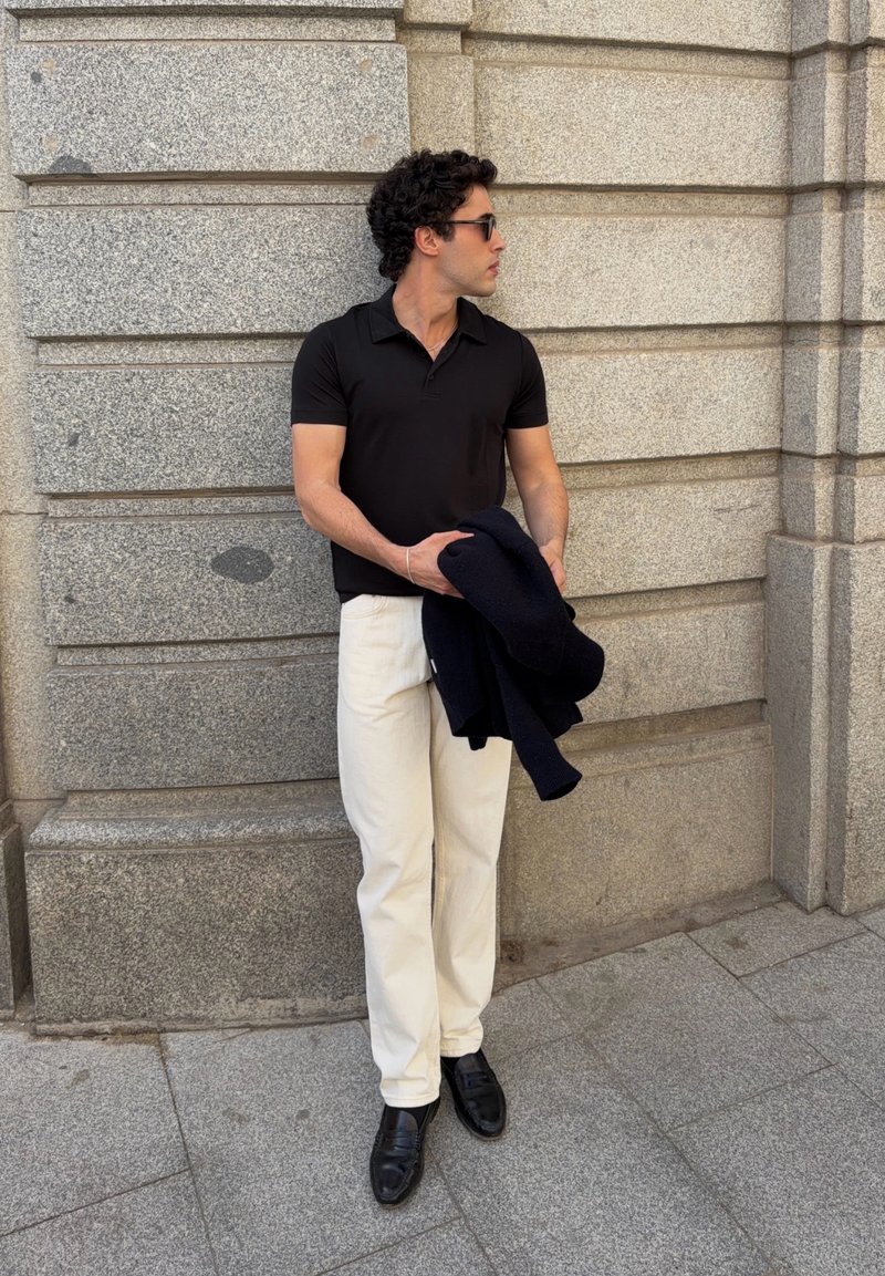 Homme portant un polo noir, un pantalon blanc et des chaussures noires, tenant une veste sombre pliée, debout contre un mur en pierre grise.