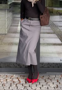 Pessoa em pé sobre um pavimento de pedra, vestindo uma blusa preta, uma saia midi cinza-clara, collants pretos, sapatos de salto vermelho com laços e carregando uma bolsa de ombro castanha.