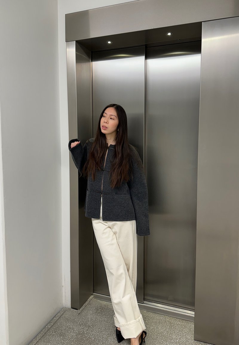 Siva jakna od mješavine vune s patentnim zatvaračem sprijeda i dugim rukavima, uparena s bijelim širokim hlačama i crnim štiklama, stoji pored metalnog lifta.