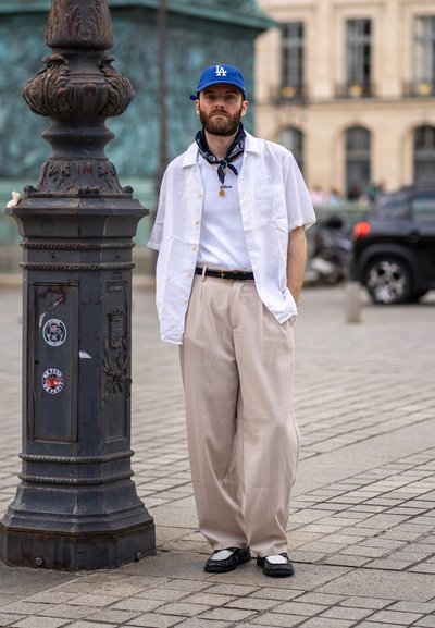 Camisa blanca de manga corta, camiseta blanca, pantalones anchos beige, zapatos negros con detalles blancos, gorra de béisbol azul y bufanda con estampado.