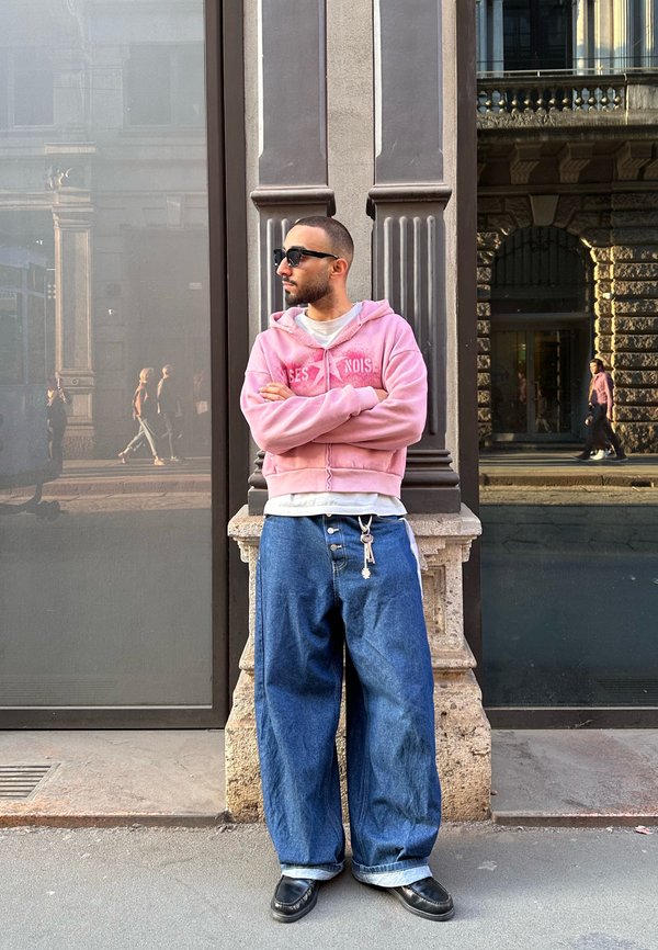 Mann mit Sonnenbrille, der einen pinken Hoodie und weite blaue Jeans trägt, lehnt sich bei Sonnenuntergang an eine verzierte Säule auf einer Stadtstraße.