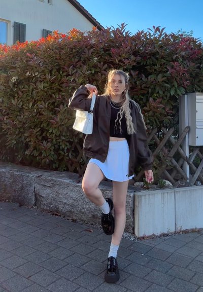 Chaqueta bomber marrón, top corto negro, falda blanca, zapatos de plataforma negros, bolso blanco. De pie en un borde de piedra con vegetación detrás.