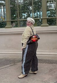 Personne portant un béret beige et une veste avec un pantalon à imprimé léopard, portant un sac à main marron avec une peluche à l'extérieur près de grandes fenêtres.