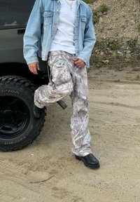 Cargo hlače s kamuflažnim uzorkom u nijansama sive i zelene, uparene s laganom traper jaknom i crnim čizmama, na pješčanoj pozadini.