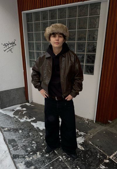 Chaqueta bomber de cuero marrón, sudadera con capucha negra, pantalones anchos negros, gorro de piel, anillos metálicos. Suelo nevado y fondo de cristal texturizado.
