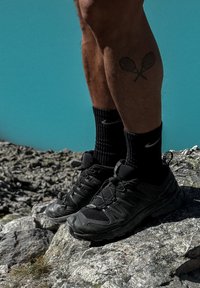 Jalad, millel on risti tätoveeritud tennisesulgpallireket, mustade sportlike sokkide ja kingade kohal, seisavad kivisel maastikul sinise tausta ees.