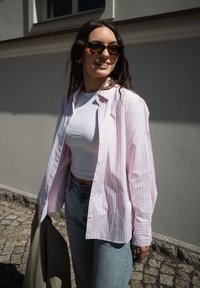 Chemise boutonnée rayée rose clair et blanche portée sur un haut blanc ajusté, assortie à un jean bleu. Des lunettes de soleil en écaille de tortue complètent la tenue.