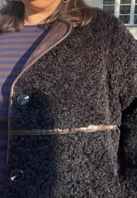 Zwarte, getextureerde jas met een zachte, pluizige stof, satijnen biezen en glanzende knopen, gedragen over een marineblauw en zwart gestreept shirt.