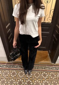 Bílé bavlněné tričko, černé sametové kalhoty s širokými nohavicemi a černá kabelka se stříbrnými kovovými detaily. Stojí na dlaždicích s vzorem.