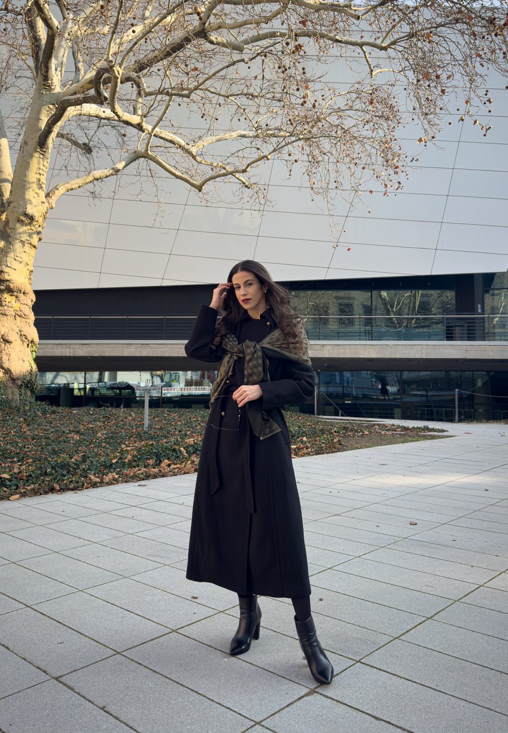 Long black coat with button details, paired with a green patterned scarf. The model wears black ankle boots, standing on a tiled surface.