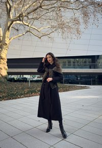 Long black coat with button details, paired with a green patterned scarf. The model wears black ankle boots, standing on a tiled surface.