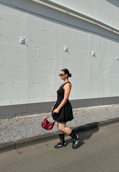Vestido negro con falda acampanada, combinado con calcetines negros y zapatillas deportivas. Sosteniendo un bolso rojo, caminando junto a una pared gris texturizada.