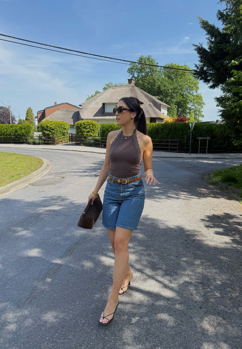 Femme portant des lunettes de soleil, un haut sans manches et un short en denim marchant dans une rue courbée près des maisons et de la verdure par une journée ensoleillée.