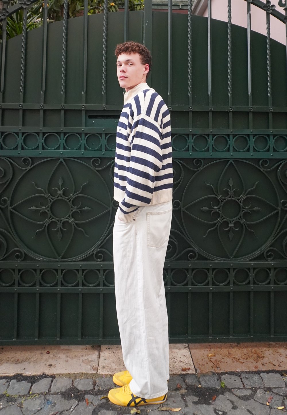 Gestreepte marineblauwe en witte trui, witte wijde broek en gele sneakers. Het model staat tegen een groene decoratieve poort.