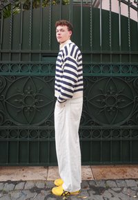 Gestreepte marineblauwe en witte trui, witte wijde broek en gele sneakers. Het model staat tegen een groene decoratieve poort.