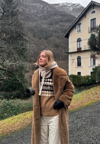 Ξανθιά γυναίκα φορώντας καφέ γούνινο παλτό, πουλόβερ με σχέδια, μπεζ κασκόλ και μαύρα γάντια, στέκεται σε ένα μονοπάτι κοντά σε ένα σπίτι σε ορεινό τοπίο.