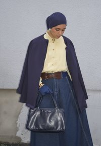 Femme portant un foulard bleu marine, une cape, des gants bleus, un chemisier jaune, une jupe en denim, tenant un sac à main noir contre un mur blanc texturé.