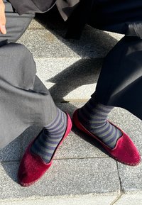 Karmīnas vilnas čības ar noapaļotu purngali un melnu apdari, kopā ar tumši zilu un zaļu svītru zeķēm, uz pelēku flīžu virsmas.