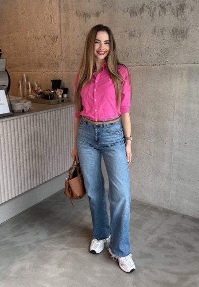 Camisa rosa de botones, vaqueros acampanados azul claro, zapatillas blancas y un bolso marrón. Fondo de hormigón liso con decoración moderna.