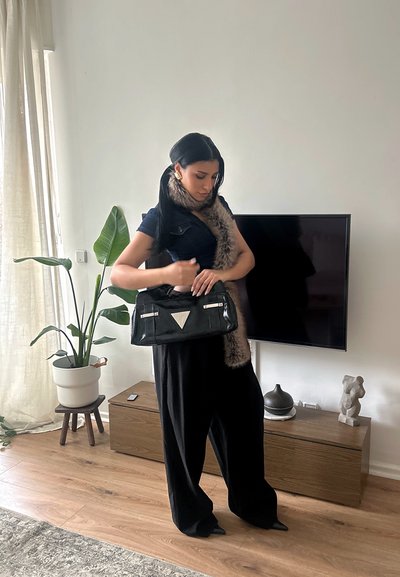 Mujer de cabello largo y oscuro, con una bufanda de piel y un atuendo negro, de pie en el interior sosteniendo un bolso negro cerca de un televisor y un armario de madera.