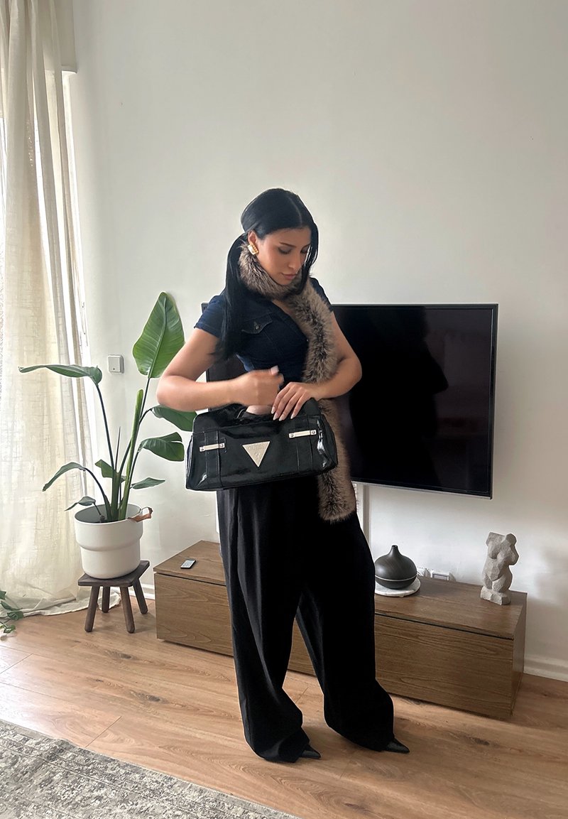 Femme aux longs cheveux noirs portant une écharpe en fourrure et une tenue noire, debout à l'intérieur, tenant un sac à main noir près d'une télévision et d'une armoire en bois.