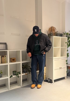 Chaqueta bomber negra sobre una sudadera con capucha verde, vaqueros azules y zuecos marrones. Estanterías con libros y plantas en un interior minimalista.