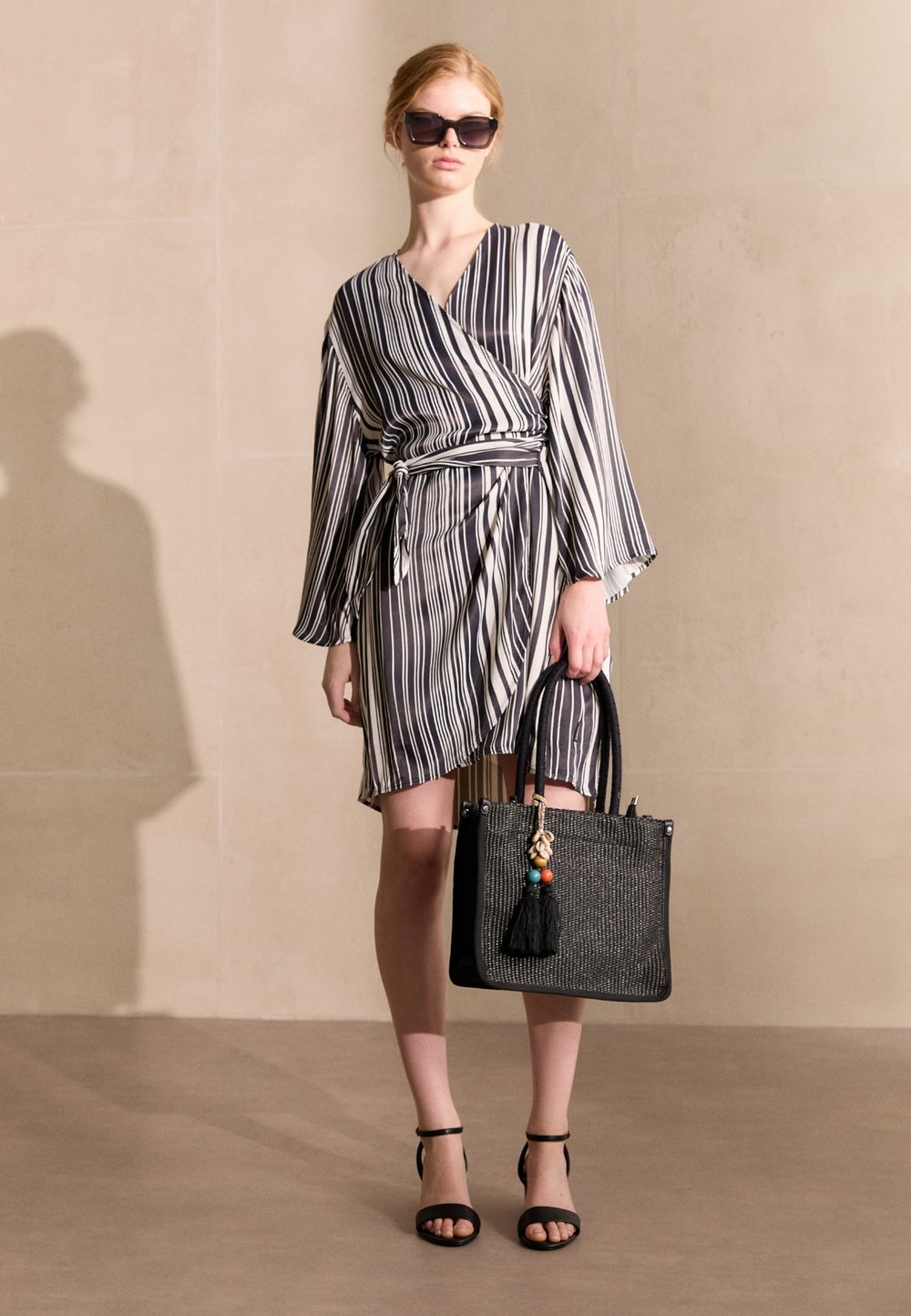 Zwart-wit gestreepte wikkeljurk met lange klokmouwen, gecombineerd met een zwarte tas met een geweven textuur en kleurrijke franje.