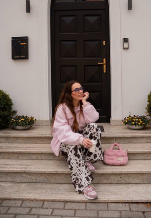 Joven mujer con chaqueta rosa y pantalones con estampado de vaca sentada en los escalones sosteniendo una taza, con un bolso rosa a su lado junto a una puerta negra.