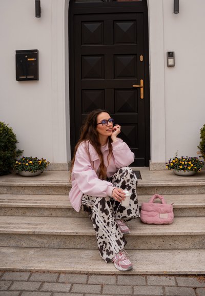 Joven mujer con chaqueta rosa y pantalones con estampado de vaca sentada en los escalones sosteniendo una taza, con un bolso rosa a su lado junto a una puerta negra.